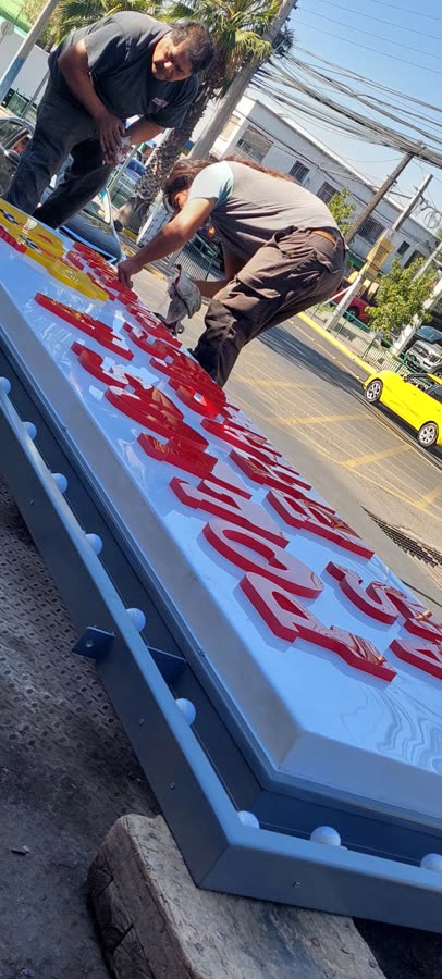 Técnicos instalando letras corpóreas rojas sobre letrero panaflex en el taller